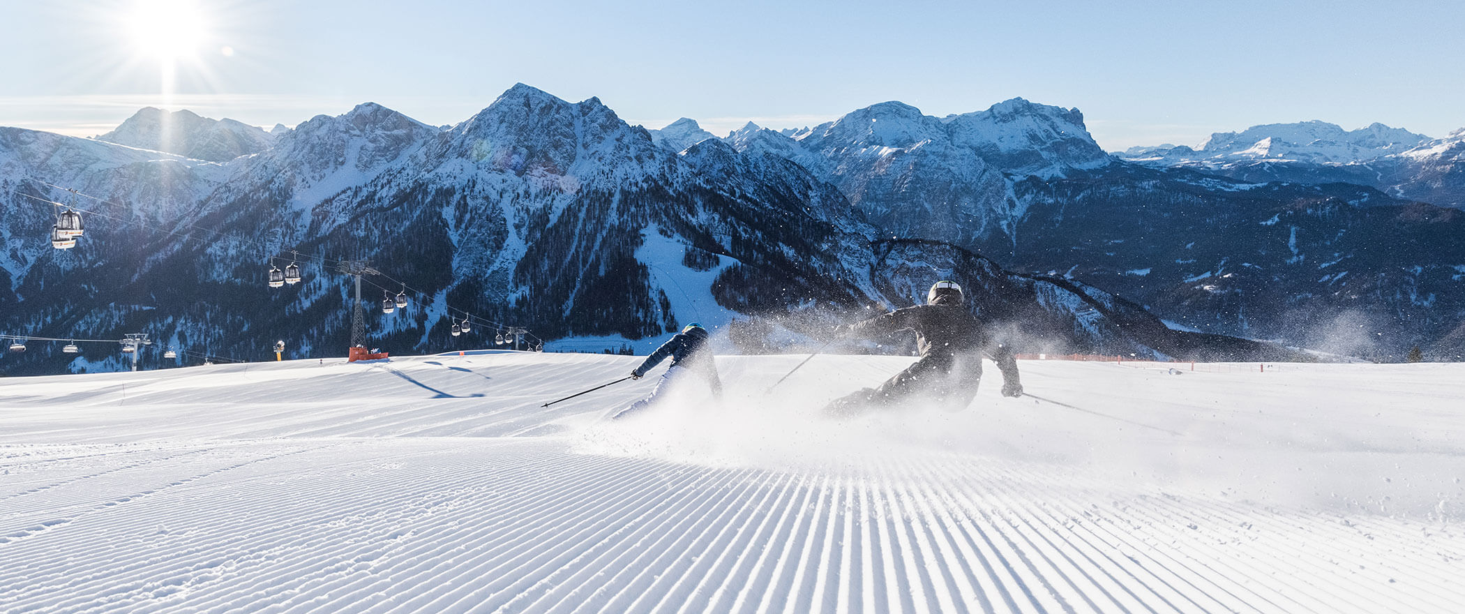 Zwei Skifahrer fahren die Piste runter, dahinter die Gondeln und die verschneiten Gipfel der Dolomiten - Hotel Majestic
