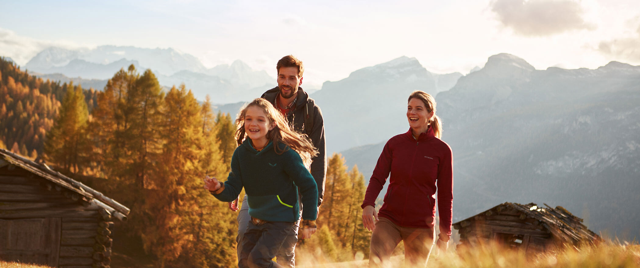 Mama, Papa und Tochter auf einer Almenwiese, dahinter zwei Almen und die bunten Herbstwälder - Hotel Majestic